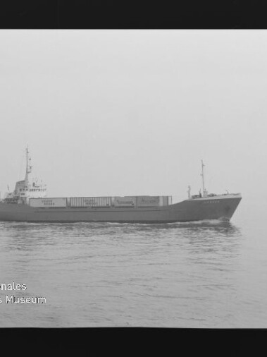Schwarzweißaufnahme eines Frachtschiffs mit Containern, das bei ruhiger, nebliger See von links nach rechts fährt; Filmrand und Wasserzeichen des Internationalen Maritimen Museums Hamburg sichtbar.