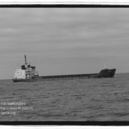 Schwarzweißaufnahme eines langen Frachtschiffs mit achtern liegenden Aufbauten, das bei ruhiger See am Horizont fährt; im linken unteren Eck das Wasserzeichen des Internationalen Maritimen Museums Hamburg.