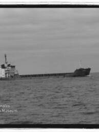 Schwarzweißaufnahme eines langen Frachtschiffs, das bei leicht bewegter See unter einem bedeckten Himmel von links nach rechts fährt; unten links sichtbar das Logo und der Schriftzug des Internationalen Maritimen Museums Hamburg.