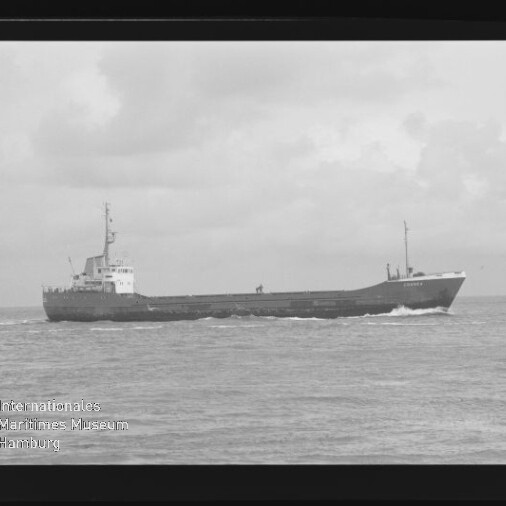 Schwarzweiß-Foto eines Frachtschiffs in Seitenansicht, das mit leichter Bugwelle über ruhige See fährt vor bewölktem Himmel; unten links ist ein Wasserzeichen des Internationalen Maritimen Museums Hamburg sichtbar.