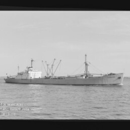 Schwarzweiß-Foto einer Frachtschiff-Seitenansicht auf ruhiger See mit sichtbaren Ladekränen und Masten, rechteckigem Filmrand und Museumslogo in der unteren linken Ecke.