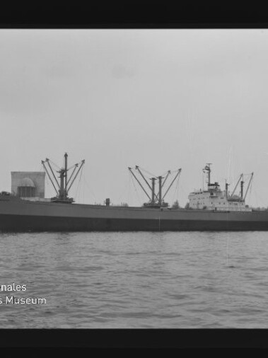 Schwarzweißaufnahme eines seitlich sichtbaren Frachtschiffs im Hafen mit mehreren Ladekränen und Lagergebäuden im Hintergrund, Wasser im Vordergrund und dem Logo des Internationalen Maritimen Museums Hamburg unten links.