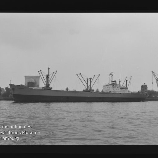 Schwarzweißaufnahme eines seitlich sichtbaren Frachtschiffs im Hafen mit mehreren Ladekränen und Lagergebäuden im Hintergrund, Wasser im Vordergrund und dem Logo des Internationalen Maritimen Museums Hamburg unten links.