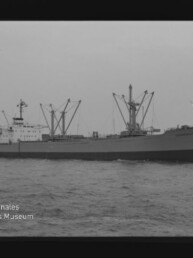 Schwarzweiß-Fotografie eines seitenansichtigen Frachtschiffs mit mehreren Ladebäumen und Kranen, fahrend vor einem Hafen mit weiteren Hafenkränen, Filmrand sichtbar und Museumswasserzeichen unten links.