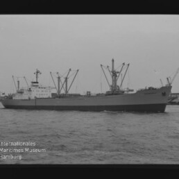 Schwarzweiß-Fotografie eines seitenansichtigen Frachtschiffs mit mehreren Ladebäumen und Kranen, fahrend vor einem Hafen mit weiteren Hafenkränen, Filmrand sichtbar und Museumswasserzeichen unten links.