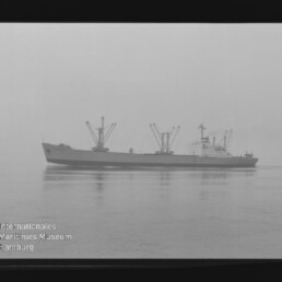 Graustufige Aufnahme eines Frachtschiffs mit mehreren Ladekränen, ruhig auf spiegelnder, diesiger See; links unten Wasserzeichen des Internationalen Maritimen Museums Hamburg und sichtbarer schwarzer Negativrand.
