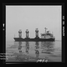 Schwarzweißaufnahme eines Frachtschiffs mit drei großen Ladekranen, seitlich auf ruhigem, spiegelndem Wasser; Filmrand mit Aufdruck 'AGFAPAN 100 356' und Logo des Internationalen Maritimen Museums Hamburg in der Ecke.