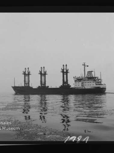 Schwarzweißaufnahme eines Frachtschiffs mit drei großen Ladekranen, seitlich auf ruhigem, spiegelndem Wasser; Filmrand mit Aufdruck 'AGFAPAN 100 356' und Logo des Internationalen Maritimen Museums Hamburg in der Ecke.