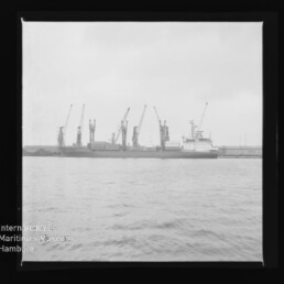 Blick über ruhiges Wasser auf ein angelegtes Frachtschiff am Kai vor einer Reihe hoher Hafenkräne unter bedecktem Himmel, mit Museumswasserzeichen unten links.