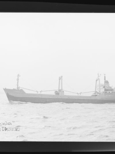 Schwarzweißaufnahme eines Frachtschiffs mit mehreren Ladekränen und Aufbauten am Heck, das auf ruhiger, leicht welliger See nahe dem Horizont fährt; unten links ist ein Museumsstempel sichtbar.