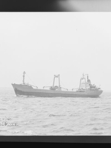 Schwarzweißaufnahme eines Frachtschiffs auf leichter See; links ein kleines Boot sowie sichtbarer Filmnegativrand und Museumslogo unten links.
