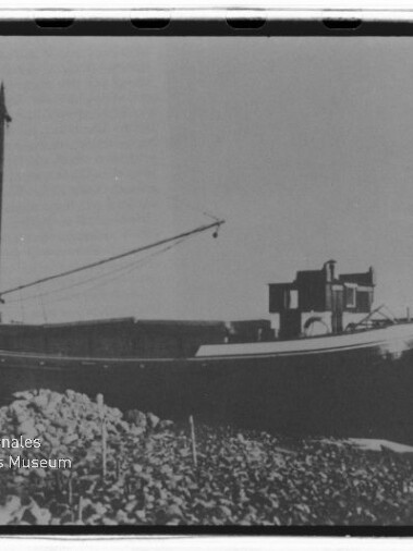 Schwarzweißaufnahme eines auf felsiger Küste aufgelaufenen Frachtschiffs mit hohem Mast und Ausleger, flachem Rumpf und kleinem Aufbau, ruhiges Meer im Hintergrund.