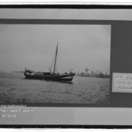 Schwarzweißaufnahme eines einmastigen Segelboots auf ruhigem Wasser vor einem Industriehafen mit Kränen; das Bild ist in einem Passepartout montiert, rechts ein handschriftliches Etikett und unten links ein Museumslogo.