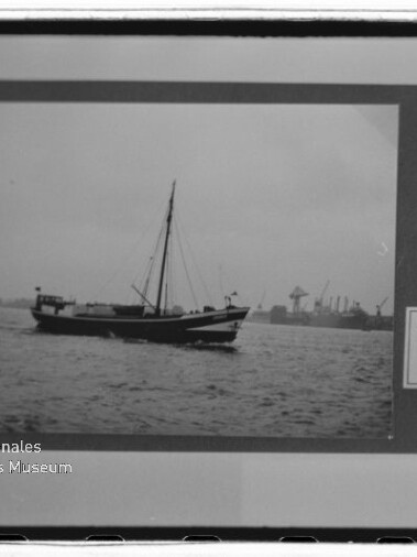 Schwarzweißaufnahme eines einmastigen Segelboots auf ruhigem Wasser vor einem Industriehafen mit Kränen; das Bild ist in einem Passepartout montiert, rechts ein handschriftliches Etikett und unten links ein Museumslogo.