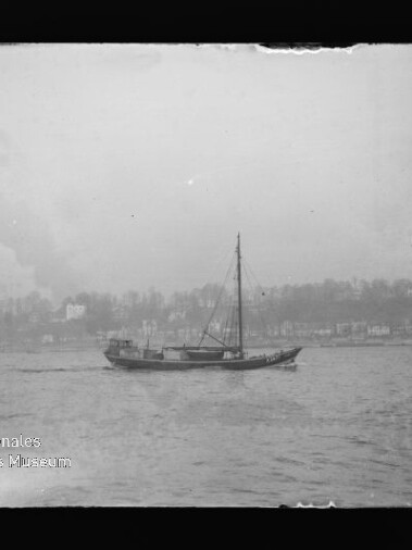 Schwarzweißfoto einer einmastigen Segelyacht auf ruhigem Wasser vor einer Uferlinie mit Häusern und Bäumen; am unteren linken Bildrand sichtbares Logo des Internationalen Maritimen Museums Hamburg.