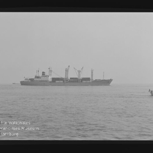 Schwarzweißaufnahme eines Frachtschiffs in ruhiger See, rechts ein kleines Beiboot mit Personen, links unten sichtbarer Filmrand und Wasserzeichen des Internationalen Maritimen Museums Hamburg.