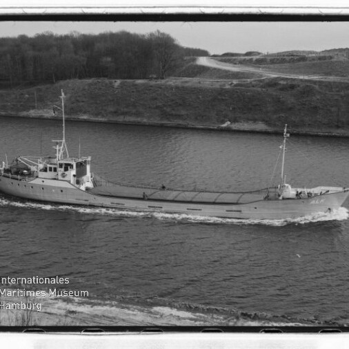 Schwarzweißfoto eines kleinen Frachtschiffs, das in einem Kanal vor einem bewaldeten Ufer entlangfährt; mittschiffs liegende Aufbauten, sichtbare Bugwelle und Museumswasserzeichen unten links.