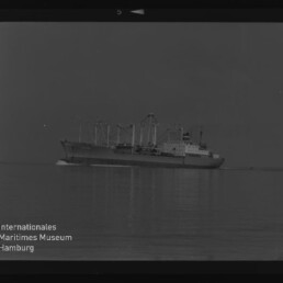 Schwarzweißaufnahme eines großen Frachtschiffs in Seitenansicht mit mehreren Ladekränen und Aufbauten, fahrend über ruhiges, spiegelndes Meer mit leichter Bugwelle; Museumswasserzeichen unten links.