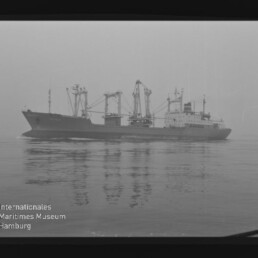 Schwarzweiß-Foto eines Frachtschiffs mit mehreren Ladekränen, das ruhig über spiegelglattes Wasser fährt, mit sichtbarer Spiegelung und dem Schriftzug „Internationales Maritimes Museum Hamburg“ unten links.