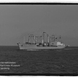 Schwarzweiß-Foto eines Frachtschiffs mit mehreren Ladekränen, das bei ruhiger See auf dem Meer fährt; unten links Logo und Schriftzug des Internationalen Maritimen Museums Hamburg.