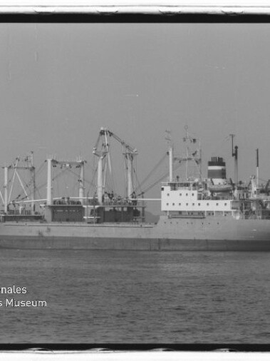 Seitliche Schwarz-Weiß-Aufnahme eines Frachtschiffs mit mehreren Ladekränen, markantem Aufbau und einem Schornstein, ruhig auf dem Meer; Wasserzeichen des Internationalen Maritimen Museums Hamburg unten links.