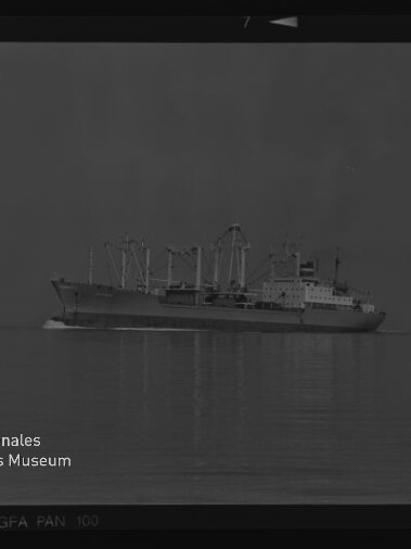 Schwarzweiß-Foto eines Frachtschiffs mit mehreren Ladekränen, das ruhig über spiegelglatte See fährt; Negativrand und Signet des Internationalen Maritimen Museums Hamburg sind sichtbar.