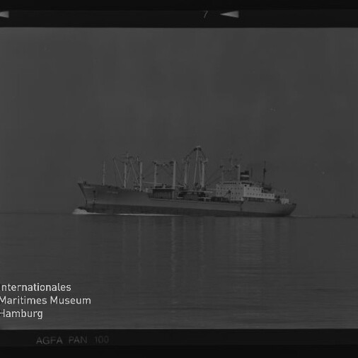 Schwarzweiß-Foto eines Frachtschiffs mit mehreren Ladekränen, das ruhig über spiegelglatte See fährt; Negativrand und Signet des Internationalen Maritimen Museums Hamburg sind sichtbar.