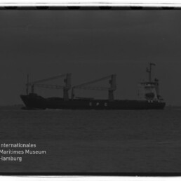 Seitliche Aufnahme eines Frachtschiffs mit zwei Ladekränen und den Buchstaben CPC am Rumpf auf ruhiger See unter grauem Himmel, mit dem Wasserzeichen des Internationalen Maritimen Museums Hamburg in der Ecke.
