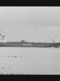 Schwarzweiß-Foto eines Frachtschiffs mit großem Schriftzug COASTAL am Rumpf, das vor flachem Küstenhintergrund auf ruhigem Wasser fährt; Filmrand und Museumslogo unten links sichtbar.