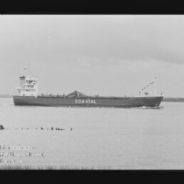 Schwarzweiß-Foto eines Frachtschiffs mit großem Schriftzug COASTAL am Rumpf, das vor flachem Küstenhintergrund auf ruhigem Wasser fährt; Filmrand und Museumslogo unten links sichtbar.