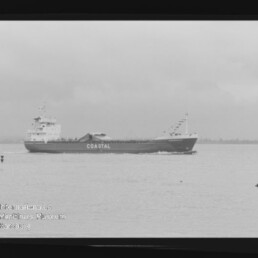 Schwarz-weiß-Foto eines mittleren Frachtschiffs mit der Aufschrift COASTAL, das auf ruhiger See parallel zur Küstenlinie fährt; im Bild sichtbarer Filmrand und Museumswasserzeichen.