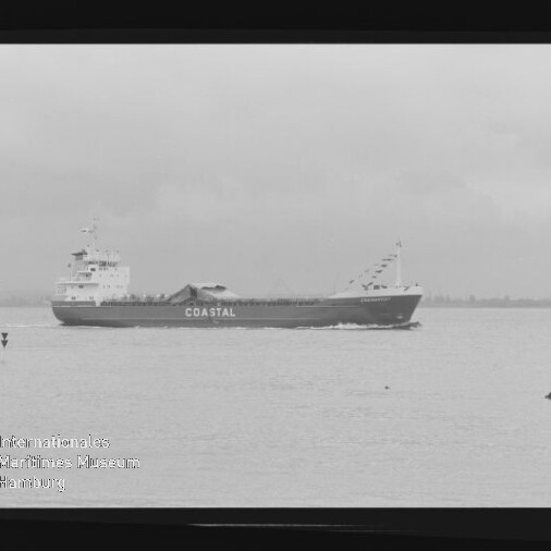 Schwarz-weiß-Foto eines mittleren Frachtschiffs mit der Aufschrift COASTAL, das auf ruhiger See parallel zur Küstenlinie fährt; im Bild sichtbarer Filmrand und Museumswasserzeichen.