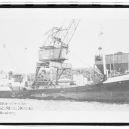Schwarz-weiß-Foto eines Frachtschiffs namens „Crail“, am Kai liegend, mit großem Hafenkran auf dem Deck und Industrieanlagen im Hintergrund.