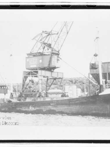 Schwarz-weiß-Foto eines Frachtschiffs namens „Crail“, am Kai liegend, mit großem Hafenkran auf dem Deck und Industrieanlagen im Hintergrund.