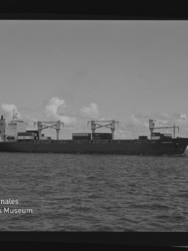 Schwarzweiß-Foto eines Frachtschiffs in ruhiger See, seitliche Ansicht mit drei sichtbaren Ladekränen und Containern auf dem Deck, flacher Horizont und vereinzelte Wolken im Hintergrund sowie Logo des Internationalen Maritimen Museums Hamburg in der linken unteren Ecke.
