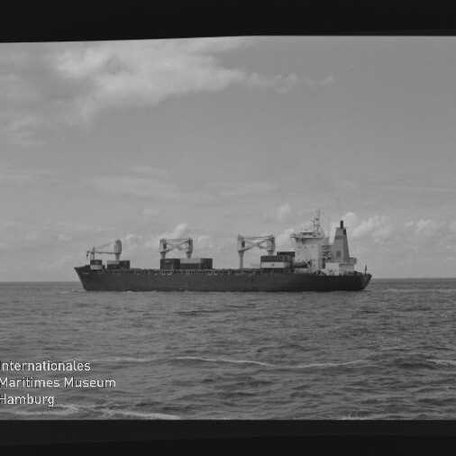 Schwarzweißaufnahme eines seitlich fotografierten Frachtschiffs mit mehreren Ladekränen auf offener See unter leicht bewölktem Himmel; links unten das Wasserzeichen des Internationalen Maritimen Museums Hamburg.