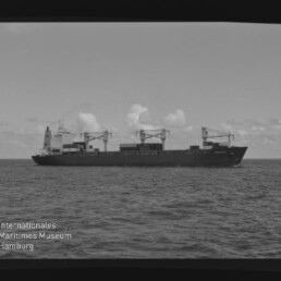 Schwarzweiß-Aufnahme eines Frachtschiffs mit drei Ladekränen und Containerdeck, das in ruhiger See unter leicht bewölktem Himmel fährt; unten links das Logo 