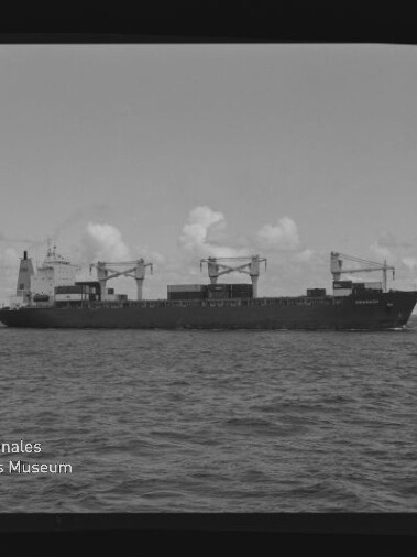 Schwarzweiß-Aufnahme eines Frachtschiffs mit drei Ladekränen und Containerdeck, das in ruhiger See unter leicht bewölktem Himmel fährt; unten links das Logo 
