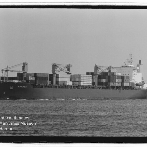 Containerschiff 'CRANACH' auf ruhiger See, beladen mit gestapelten Containern und drei Deckskränen; Schwarzweißaufnahme mit Museumswasserzeichen unten links.