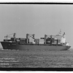 Schwarzweißaufnahme eines beladenen Containerschiffs mit gestapelten Containern und Deckskränen auf dem Meer, mit sichtbaren Filmrändern und Wasserzeichen des Internationalen Maritimen Museums Hamburg.