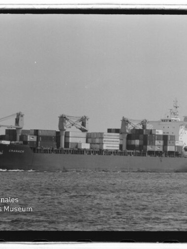 Schwarzweißaufnahme eines beladenen Containerschiffs mit gestapelten Containern und Deckskränen auf dem Meer, mit sichtbaren Filmrändern und Wasserzeichen des Internationalen Maritimen Museums Hamburg.