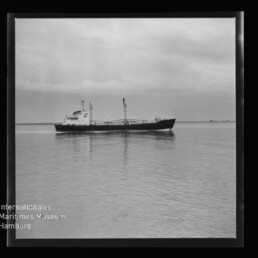 Schwarzweiß-Fotografie eines Frachters mit Aufbauten und Ladebaum, der ruhig auf spiegelglattem Wasser vor flachem Horizont fährt; unten links sichtbar das Logo des Internationalen Maritimen Museums Hamburg.