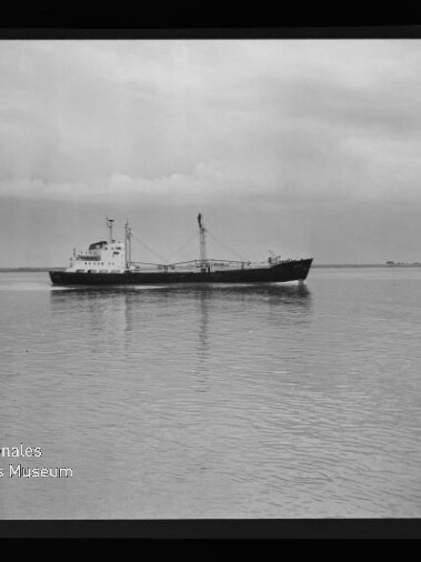 Schwarzweiß-Fotografie eines Frachters mit Aufbauten und Ladebaum, der ruhig auf spiegelglattem Wasser vor flachem Horizont fährt; unten links sichtbar das Logo des Internationalen Maritimen Museums Hamburg.