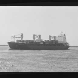Schwarzweißaufnahme eines Containerschiffs mit drei Deckskränen und gestapelten Containern, das auf ruhigem Wasser vor einer flachen Küstenlinie fährt; unten links sichtbar das Logo des Internationalen Maritimen Museums Hamburg.