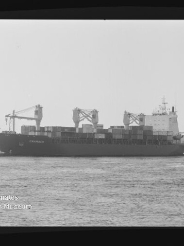 Schwarzweißaufnahme eines Containerschiffs mit drei Deckskränen und gestapelten Containern, das auf ruhigem Wasser vor einer flachen Küstenlinie fährt; unten links sichtbar das Logo des Internationalen Maritimen Museums Hamburg.