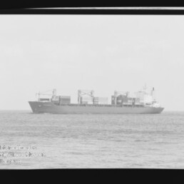 Schwarzweißaufnahme eines containerbeladenen Frachtschiffs in Seitenansicht auf ruhiger See, mit Ladekränen und gestapelten Containern, sichtbaren Filmrändern und Wasserzeichen des Internationalen Maritimen Museums Hamburg.