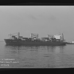 Schwarzweißaufnahme eines beladenen Containerschiffs auf ruhiger See mit gestapelten Containern, drei Ladekränen und einem kleinen Begleitschiff am Heck; links unten das Logo und der Schriftzug des Internationalen Maritimen Museums Hamburg sichtbar.