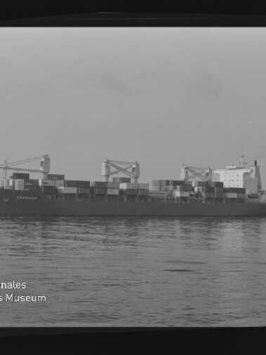 Schwarzweißaufnahme eines beladenen Containerschiffs auf ruhiger See mit gestapelten Containern, drei Ladekränen und einem kleinen Begleitschiff am Heck; links unten das Logo und der Schriftzug des Internationalen Maritimen Museums Hamburg sichtbar.