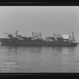 Schwarzweißaufnahme eines großen Containerschiffs mit aufgestapelten Containern und drei Ladekränen, das ruhig auf glattem Wasser vor dem Horizont fährt; links unten ist das Logo des Internationalen Maritimen Museums Hamburg zu sehen.