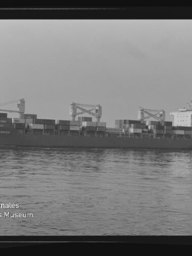 Schwarzweißaufnahme eines großen Containerschiffs mit aufgestapelten Containern und drei Ladekränen, das ruhig auf glattem Wasser vor dem Horizont fährt; links unten ist das Logo des Internationalen Maritimen Museums Hamburg zu sehen.
